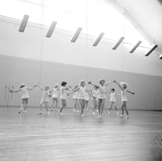 Schwarzes und weißes Foto von jungen Mädchen, die sich an den Händen halten und in einem Studio tanzen, mit einem Ball auf der rechten Seite, Deckenleuchten und einem wandmontierten Objekt im Hintergrund.