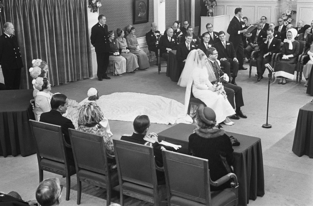 Schwarz-weiß-Foto einer Hochzeitszeremonie mit sitzenden und stehenden Menschen, einem Mann mit Mikrofon, einer Frau mit einem Blumenstrauß, einem Podium mit Mikrofon, Blumenarrangements, einem Vorhang, einem gerahmten Foto an der Wand und einer Tür im Hintergrund.
