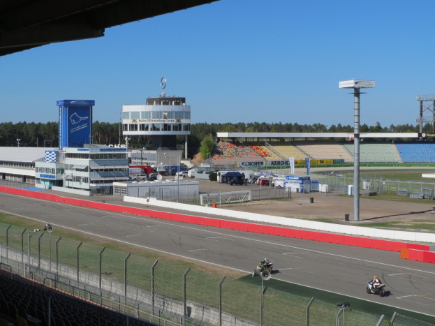 Gruppe von Motorradfahrern auf einer Rennstrecke, umgeben von einem Metallzaun, Gras, Fahrzeugen, einem Gebäude, einem Schild, Pfählen, einem Stadion, Deckenleuchten, Bäumen und einem bewölkten Himmel.