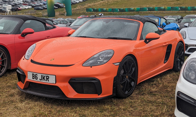 Porsche 718 Boxster GTS auf dem Goodwood Festival of Speed mit Autos auf Gras, einem Banner im Hintergrund, Bäumen und einem klaren blauen Himmel.