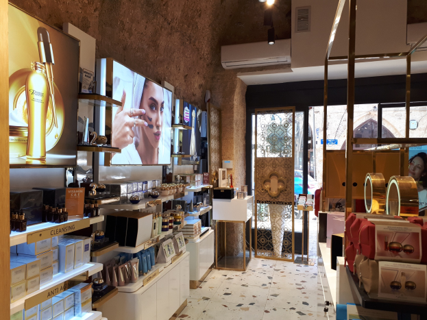 Ein Kosmetikgeschäft mit Regalen, auf denen verschiedene Schönheitsprodukte angeordnet sind, Wandbildschirme, Deckenleuchten, eine Klimaanlage und Glastüren, die eine Straßenszene in Dubai mit einer Person und einem Auto zeigen.