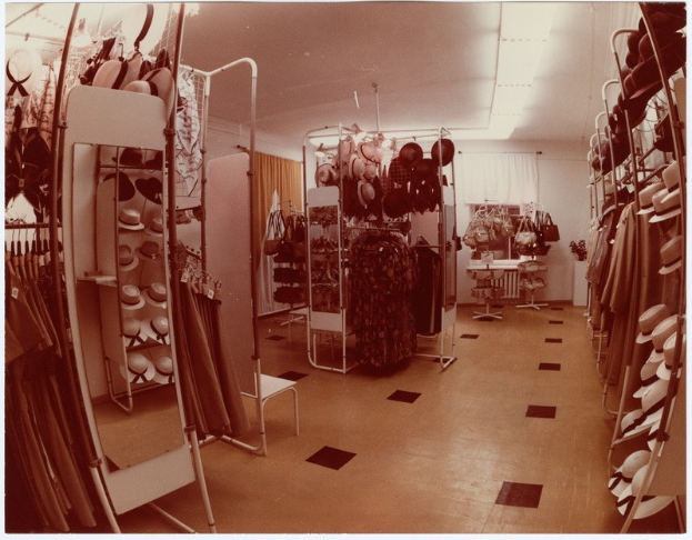 Altes Schwarz-Weiß-Foto eines kleinen Bekleidungsgeschäfts-Innenraums mit Kleiderstangen, Hut-Display, Spiegel, Stuhl, Tisch mit diversen Gegenständen, gemusterten Vorhängen und Deckenbeleuchtung.