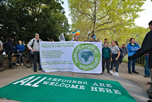 Eine Gruppe von Menschen, die draußen vor einem Banner mit der Aufschrift "Alle Flüchtlinge sind hier willkommen" und einer Fahne stehen und Fahrräder, ein Zaun, ein Straßenschild, eine Schautafel, Bäume und einen bewölkten Himmel im Hintergrund sehen.