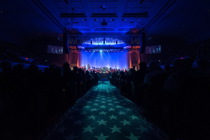 Ein großer Auditorium mit Sitzenden, eine Bühne mit Musikern und eine Decke mit Lichtern und Fernsehgeräten, was auf einen Konzertsaal für die Nationale Konferenz 2019 hindeutet.