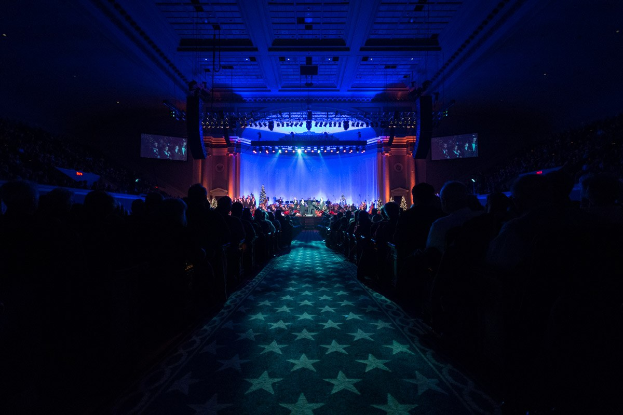Ein großer Auditorium mit Sitzenden, eine Bühne mit Musikern und eine Decke mit Lichtern und Fernsehgeräten, was auf einen Konzertsaal für die Nationale Konferenz 2019 hindeutet.