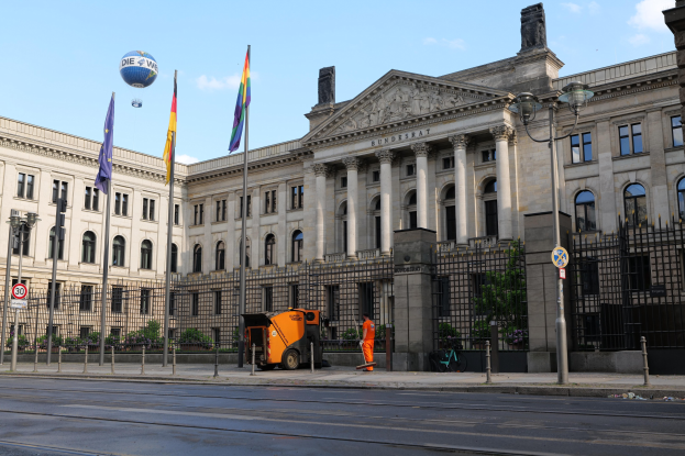 Das Reichstagsgebäude in Berlin, Deutschland, mit Flaggen, Straßenmöbeln, Fahrzeugen, Fußgängern und Bäumen vor einem bewölkten Himmel.