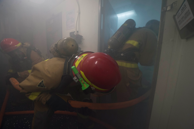 Eine Gruppe von Feuerwehrleuten in Helmen und Sauerstoffzylindern steht in einem Raum mit Rauch, der aus einer Tür kommt, mit Rohren und einer Lampe im Hintergrund und einem Objekt an der rechten Wand.