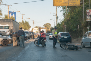 Eine Gruppe von Menschen steht um ein verunglücktes Motorrad auf der Straße herum, umgeben von mehreren Fahrzeugen, darunter ein Lastwagen, und einer Hintergrundlandschaft aus Bäumen, Pfählen, Lampen, Schildern und Himmel.