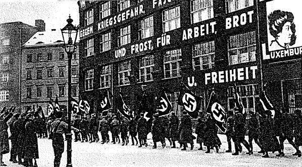 Black and white photo of a group marching down a street holding flags and banners, with a light pole on the left and a Nazi Party sign on the right.