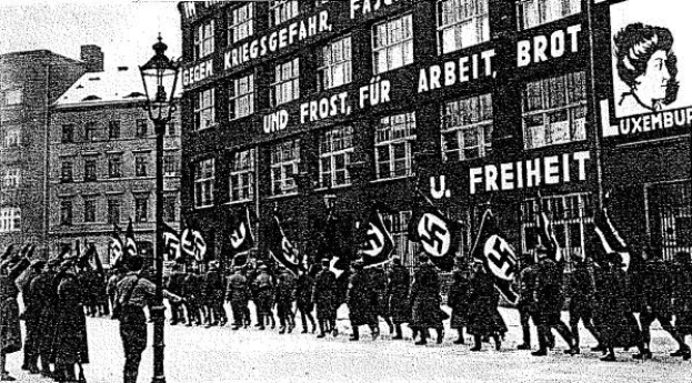 Black and white photo of a group marching down a street holding flags and banners, with a light pole on the left and a Nazi Party sign on the right.