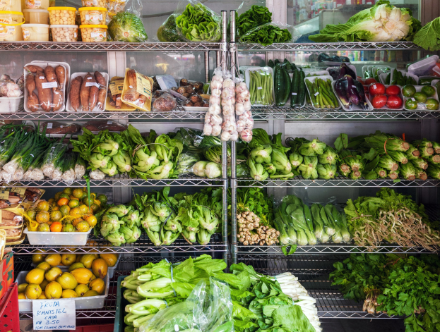 Ein Gang in einem Supermarkt mit frischem Gemüse und Obst, verpackten Artikeln in Plastikfolien, Kartons, einem Schild und einem Glasfenster im Hintergrund.