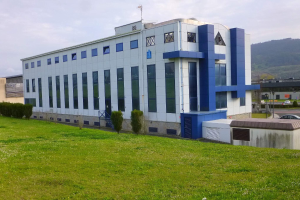 Großes weißes und blaues Gebäude mit Fahrzeugen auf der Straße, Masten, Lampen, Schildern und Bergen im Hintergrund, auf einer saftig grünen Wiese umgeben von Bäumen und Pflanzen unter einem klaren blauen Himmel.