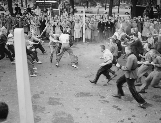 Eine Gruppe von Menschen, die um zwei Männer herumstehen, die sich vor einem Publikum körperlich streiten, mit Fahrzeugen, Bäumen, Pfählen und Gebäuden im Hintergrund, dargestellt in Schwarz-Weiß.