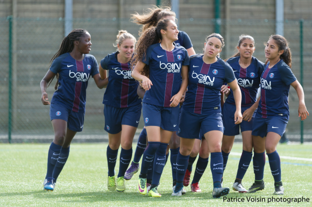 Gruppe junger Frauen, die Fußball auf einem Rasenfeld mit Maschendrahtzaun und einer Wand im Hintergrund spielen, mit der Beschriftung "Paris Saint-Germain Women's Soccer" in der unteren rechten Ecke.