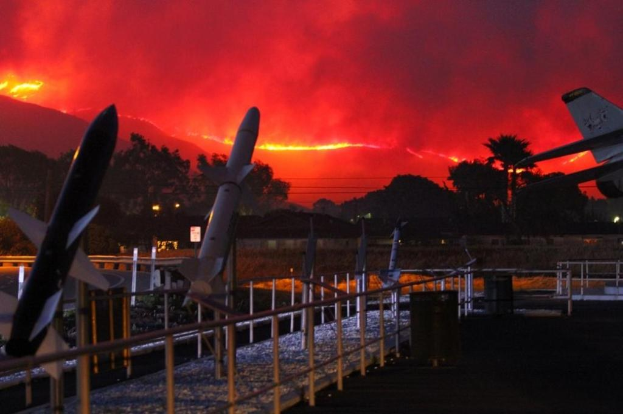 Ein Flugzeug fliegt über eine Start- und Landebahn mit Raketen darauf, umgeben von Zäunen, Mülltonnen, Bäumen, Laternenmasten und Gebäuden, mit einem von den Waldbränden in Kalifornien verqualmten Himmel im Hintergrund.