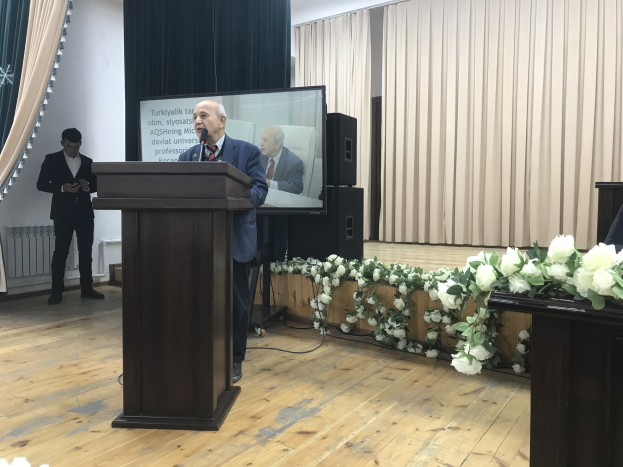 A man at a podium delivers a speech titled "Aqshining University President's Speech," with a seated person on the right and a standing person on the left, set against curtains and a wall with flowers and leaves at the bottom.