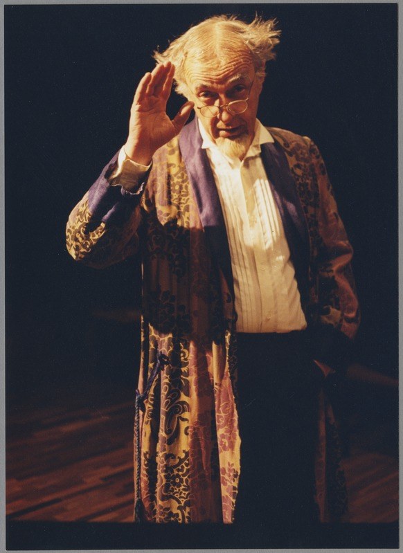 A man in a long robe and glasses stands on a stage with a dark background, clasping his hands and gazing toward the audience with a serious expression.