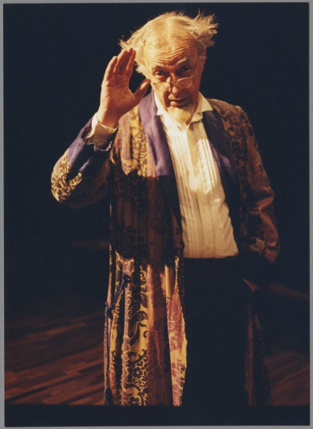A man in a long robe and glasses stands on a stage with a dark background, clasping his hands and gazing toward the audience with a serious expression.