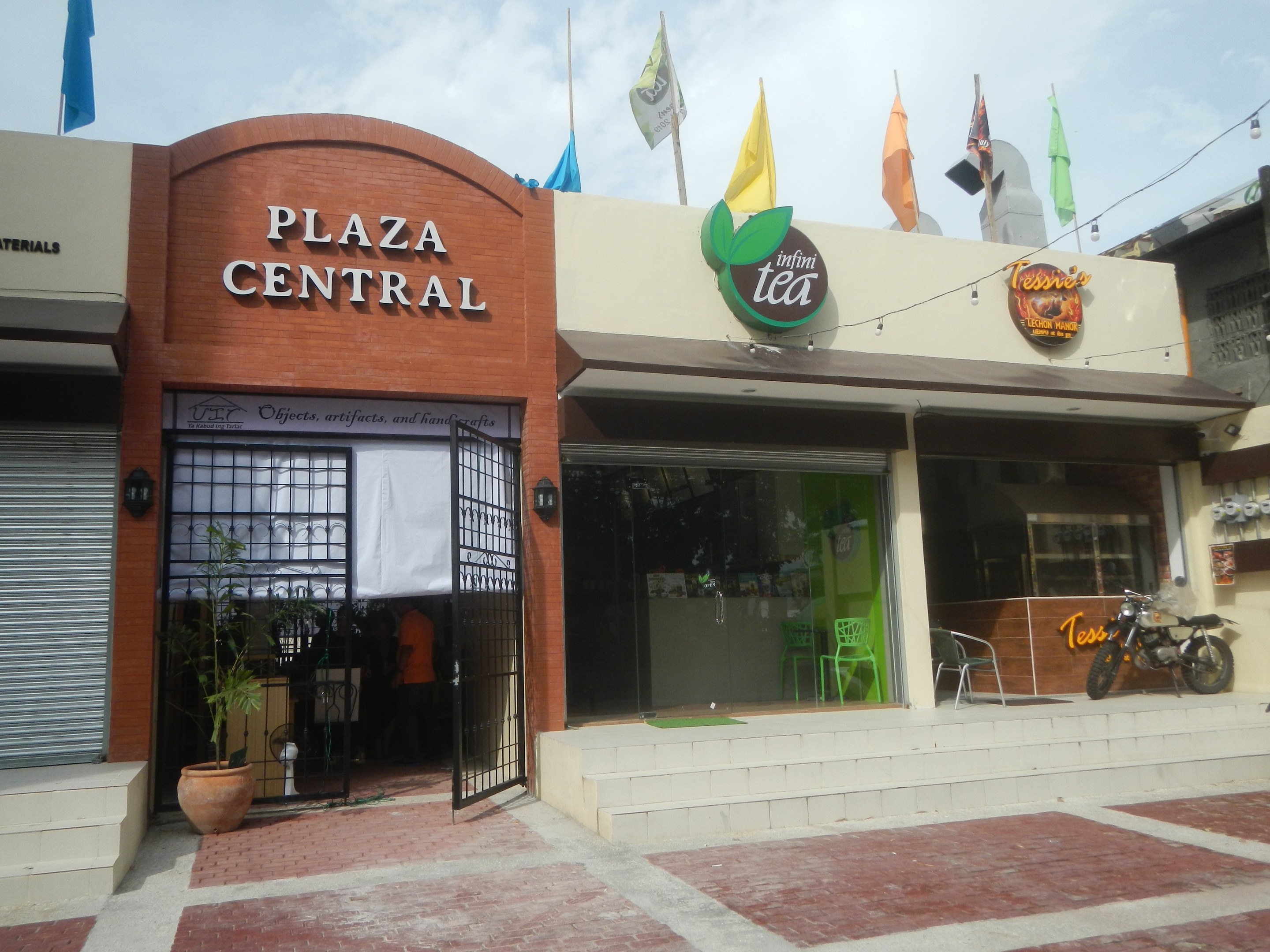 A central plaza in Cebu City, Philippines, with buildings, motorbikes, outdoor seating, greenery, flags, signage, overhead lighting, and cables against a cloudy sky.