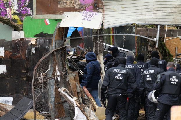 Group of police officers in black uniforms and some with helmets standing in front of a destroyed building surrounded by a metal fence, with scattered debris including wood and other objects.