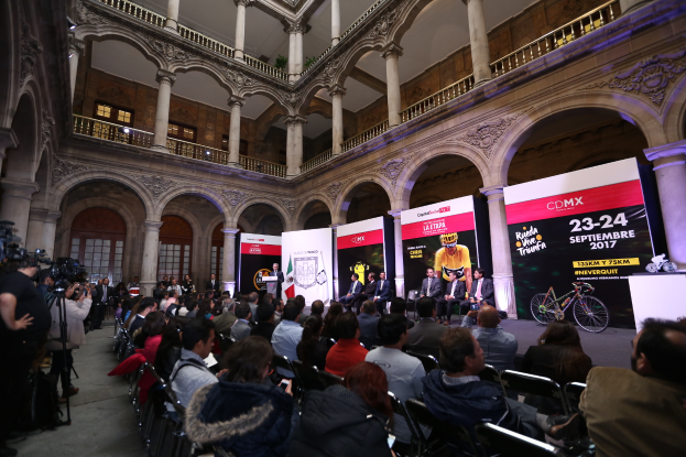 Eine Gruppe von Menschen sitzt an einem Tisch mit einem Podium und einem Mikrofon vor einer Menge, Kameras und Transparenten während einer Pressekonferenz für das CDMX 2017-Event.