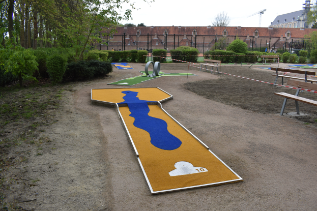 Ein Minigolfplatz in einem Park, umgeben von Bänken, Pflanzen, Bäumen und einem Zaun, mit Gebäuden und einem klaren blauen Himmel im Hintergrund.