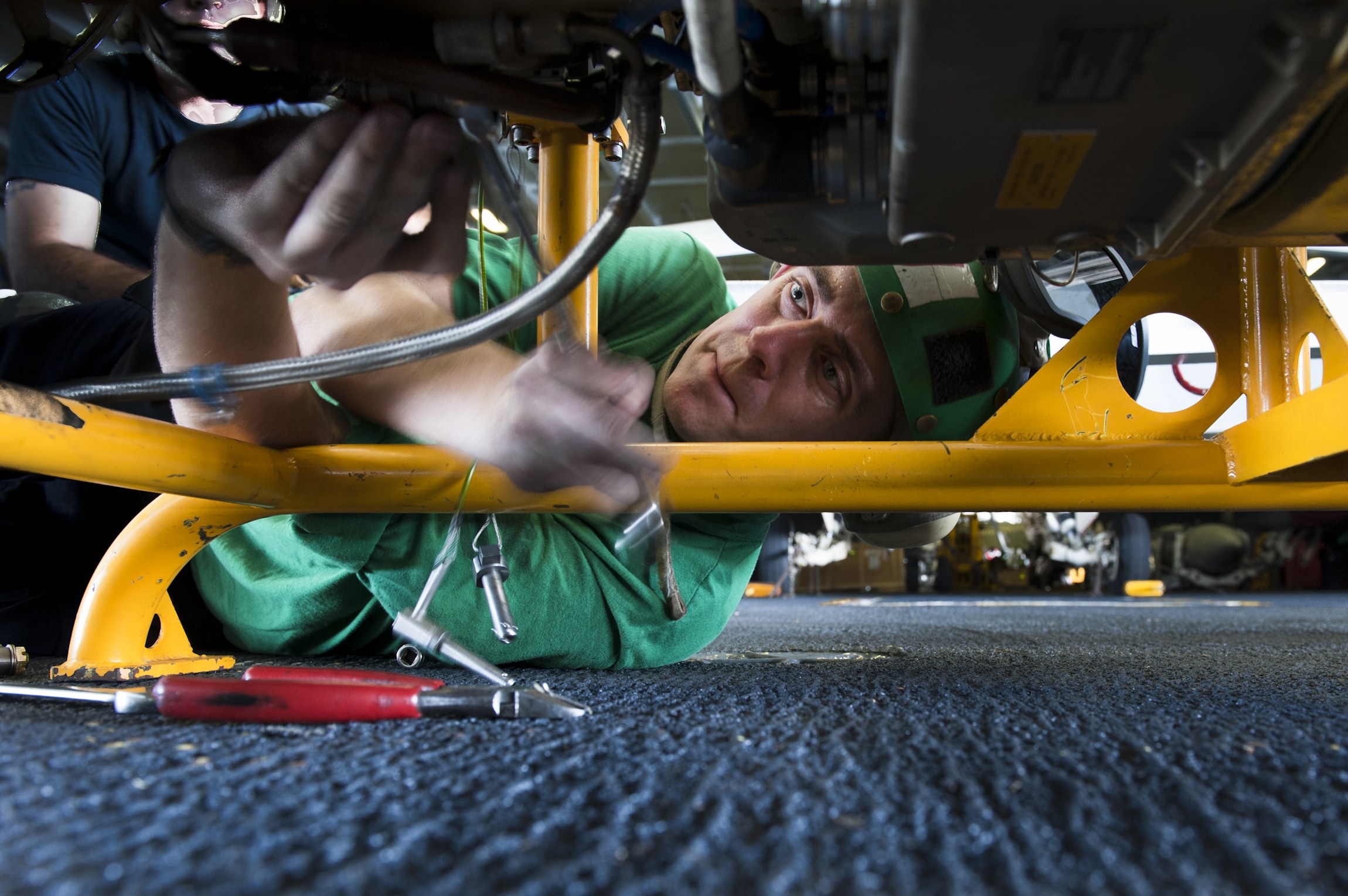 Zwei Männer arbeiten an einem Auto in einer Garage, einer liegt auf dem Boden in einem grünen T-Shirt und der andere steht hinter ihm, mit Werkzeugen drumherum.