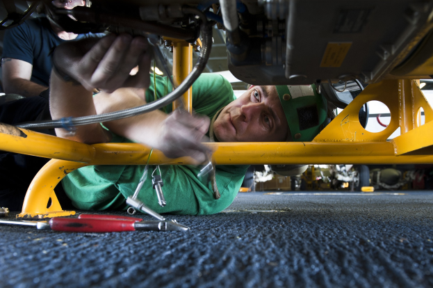 Zwei Männer arbeiten an einem Auto in einer Garage, einer liegt auf dem Boden in einem grünen T-Shirt und der andere steht hinter ihm, mit Werkzeugen drumherum.