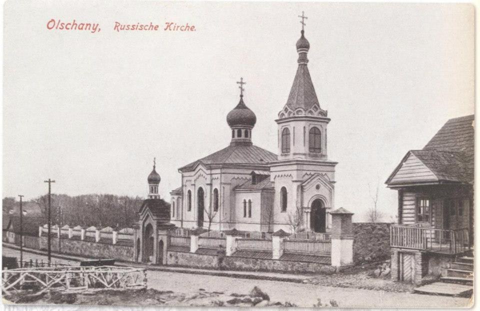 Schwarzes und weißes Foto einer russisch-orthodoxen Kirche in Olschany, Russland, umgeben von Häusern, Bäumen, Strommasten und einem Zaun unter einem klaren Himmel, mit Text oben.