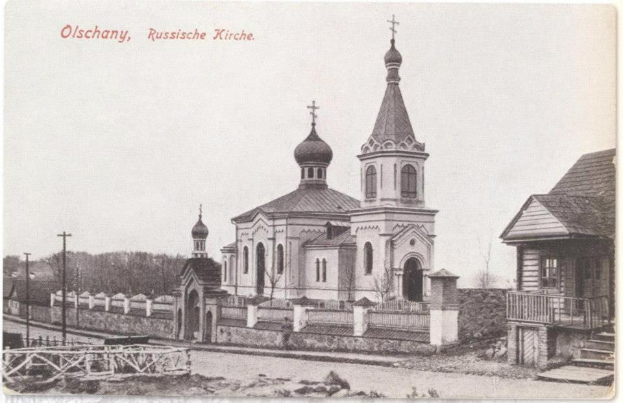Schwarzes und weißes Foto einer russisch-orthodoxen Kirche in Olschany, Russland, umgeben von Häusern, Bäumen, Strommasten und einem Zaun unter einem klaren Himmel, mit Text oben.