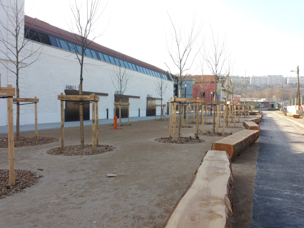 Ein Spielplatz mit Holzliegen und Bäumen vor einem Gebäude, umgeben von Straßenlaternen, Autos, Bäumen und Gebäuden unter einem klaren blauen Himmel.