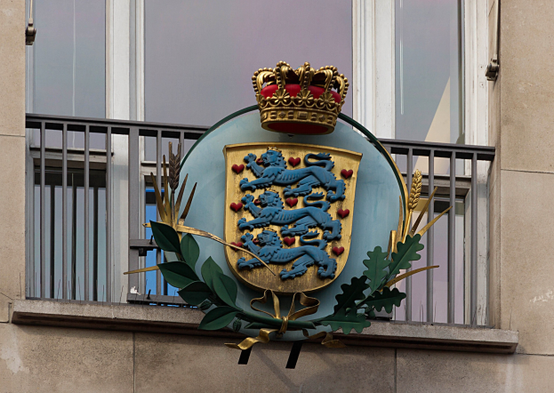 Wappen der britischen Königsfamilie, das auf der Fassade eines Gebäudes dargestellt ist, begleitet von einem Logo mit Krone und Blättern sowie Fenstern und Gittern im Hintergrund.