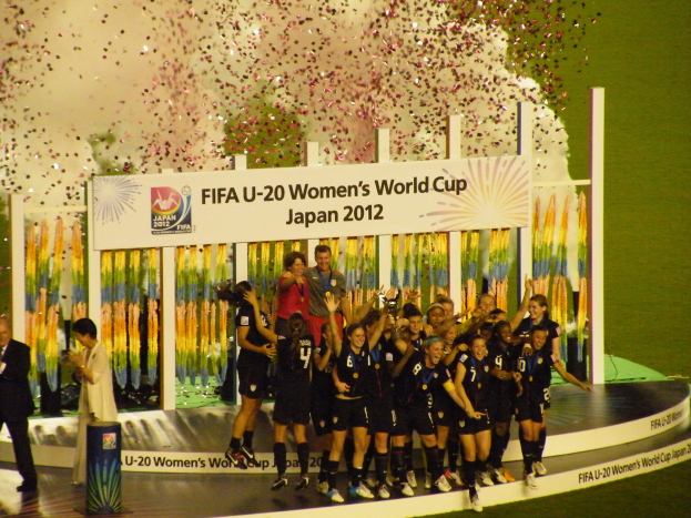 Gruppe von Frauenfußballspielerinnen in schwarzer Uniform, die auf einer Bühne umgeben von einer Menge feiern, mit einer Tafel und dekorativen Elementen im Hintergrund.