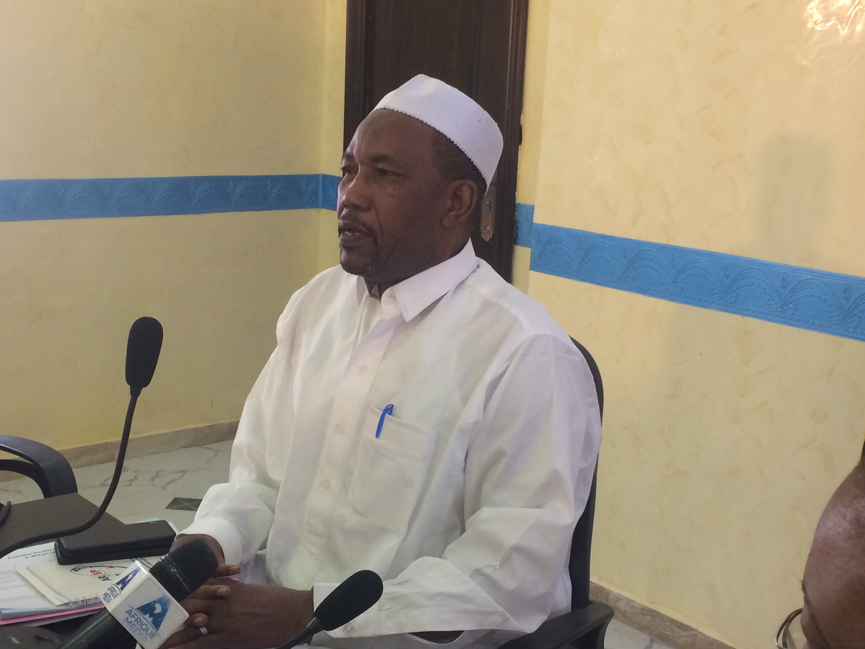 Ein Mann in einem weißen Gewand und Hut sitzt an einem Tisch mit Mikrofonen, spricht, während Papiere und Gegenstände auf dem Tisch zu sehen sind, mit einem anderen Menschen mit Brille rechts von ihm, in einem Raum mit einer Wand und einer Tür im Hintergrund.