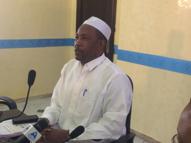 Ein Mann in einem weißen Gewand und Hut sitzt an einem Tisch mit Mikrofonen, spricht, während Papiere und Gegenstände auf dem Tisch zu sehen sind, mit einem anderen Menschen mit Brille rechts von ihm, in einem Raum mit einer Wand und einer Tür im Hintergrund.