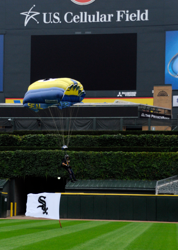 Eine Person mit einem Fallschirm über einem Baseballfeld, umgeben von grünem Gras und Pflanzen, mit einem Banner und Tafeln mit Text und Logos im Hintergrund.