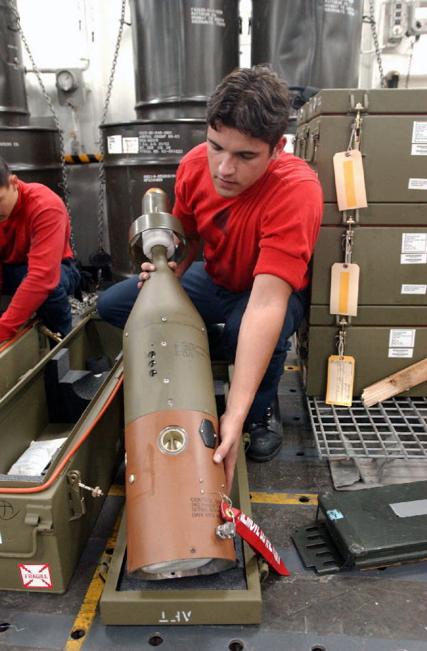 Zwei Männer in roten Hemden arbeiten an einer großen Metallfragmentierungsbombe, mit Kisten, Ketten und anderen Gegenständen im Hintergrund.