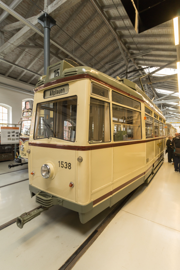 Ein gelb-cremefarbener Zug auf Eisenbahnschienen innerhalb eines Geb├Ąudes, mit einigen Menschen rechts daneben, einer Wand, einem Fenster und Deckenleuchten im Hintergrund.