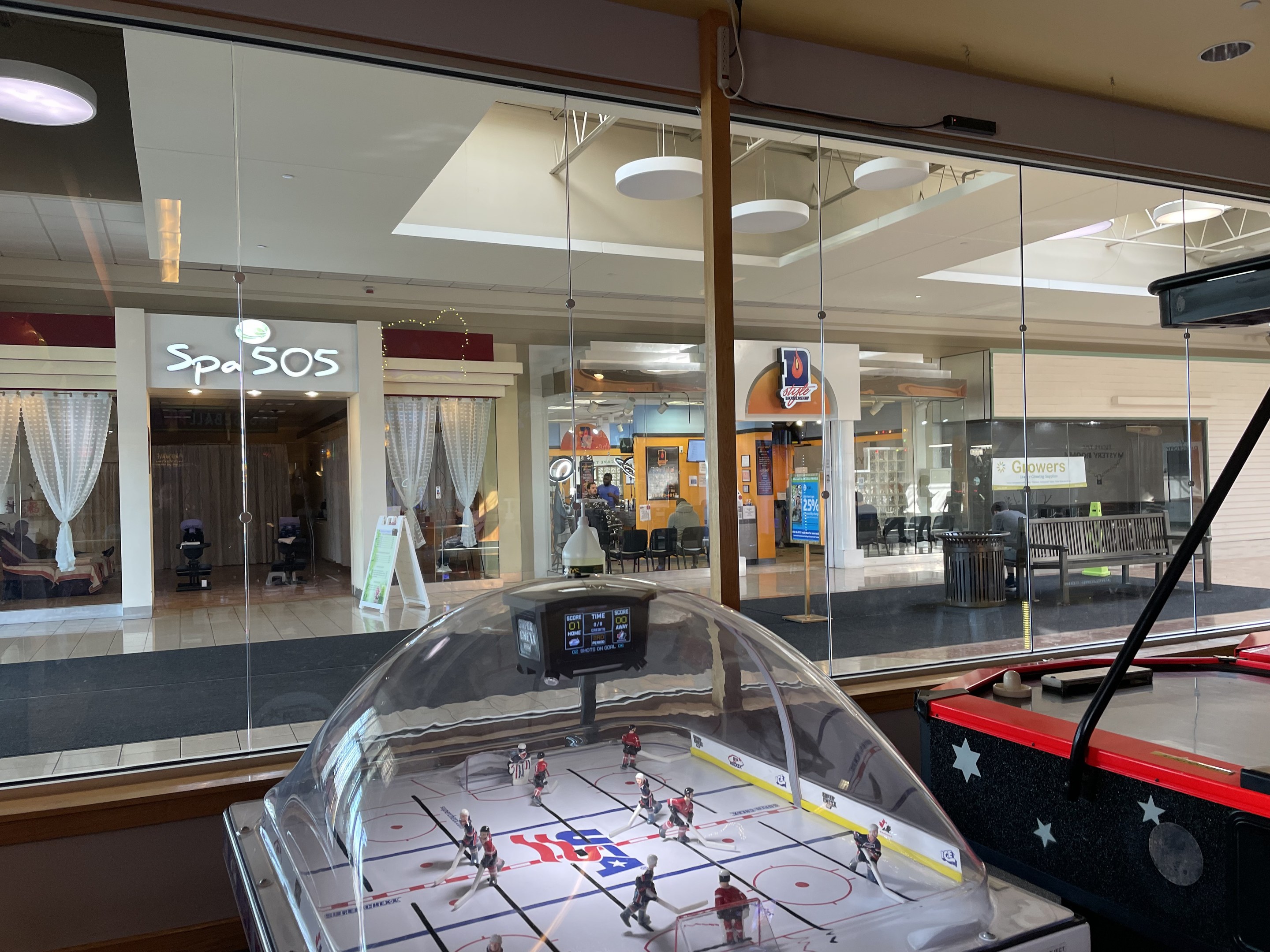 Eine Eishalle in einem Einkaufszentrum, umgeben von Glaswänden mit Menschen, Möbeln und Schildern, die von außen sichtbar sind, und einem Air-Hockey-Tisch auf der rechten Seite.