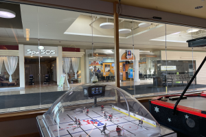 Eine Eishalle in einem Einkaufszentrum, umgeben von Glaswänden mit Menschen, Möbeln und Schildern, die von außen sichtbar sind, und einem Air-Hockey-Tisch auf der rechten Seite.