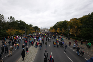 Eine große Gruppe von Menschen marschiert auf einer von Bäumen gesäumten Straße in Berlin, einige halten Kameras, mit einem Gebäude im Hintergrund und einem klaren Himmel darüber.