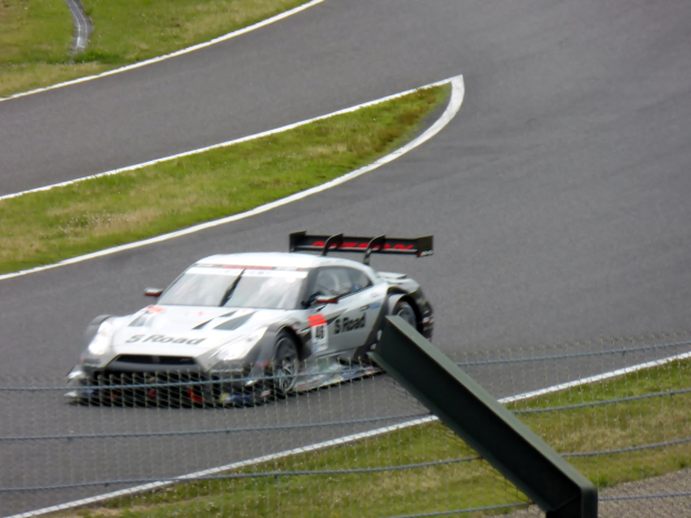 Ein Audi R8 LMS GT3-Rennwagen ist auf der Nürburgring 24 Stunden-Rennstrecke positioniert, umgeben von einem Metallzaun und saftigem Grün, mit der Strecke, die sich vor ihm erstreckt.