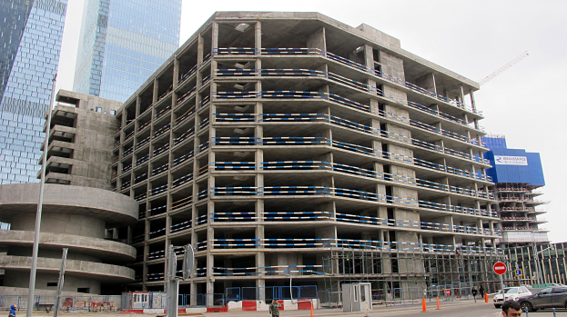 Großes Bauwerk im Bau in einer Stadt umgeben von Wolkenkratzern, mit Fahrzeugen, Fußgängern, Verkehrsinfrastruktur und Kränen vor einem klaren Himmel.