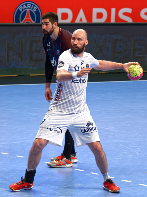 Zwei Männer spielen Handball auf einem Platz, einer hält den Ball, mit einer Tafel im Hintergrund, auf der "Paris Saint-Germain vs Paris Saint-Germain" steht.