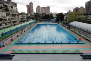 Großer städtischer Schwimmbadkomplex umgeben von Geländern, Pfählen, Laternen, Bäumen und Gebäuden, mit einem Schuppen auf der rechten Seite und Himmel im Hintergrund.