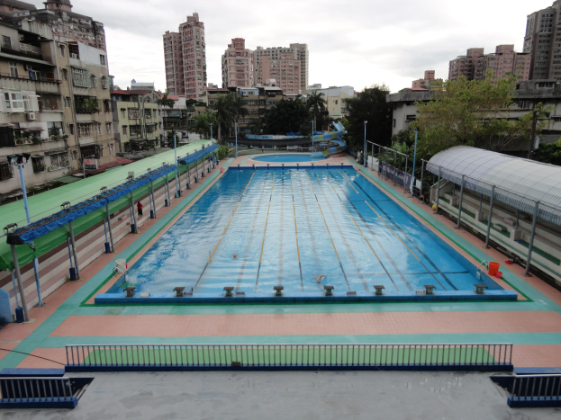 Großer städtischer Schwimmbadkomplex umgeben von Geländern, Pfählen, Laternen, Bäumen und Gebäuden, mit einem Schuppen auf der rechten Seite und Himmel im Hintergrund.